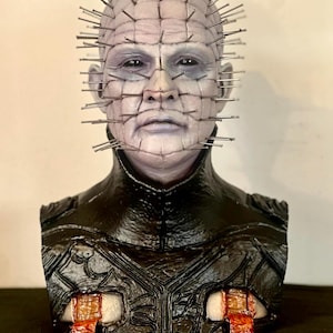 May include: A bust of a character with a pale complexion, adorned with numerous metal pins protruding from the head. The figure has a stern expression and is dressed in a black, textured garment. The bust has a horror theme.
