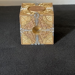 May include: A gold and silver colored puzzle box featuring intricate geometric engravings on each face. The cube has a circular indentation in the center of the front panel and a circular plate on the top panel, with metallic detailing throughout.