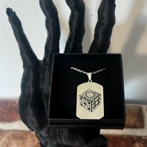 May include: A silver-colored pendant necklace featuring a detailed graphic of a puzzle box. The pendant is rectangular with a chain. The necklace is displayed in a black box, held by a black hand sculpture.