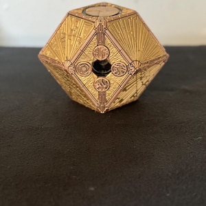 May include: A metallic gold-colored puzzle box with a complex geometric shape. The surface features intricate engraved patterns, including sunburst lines and circular symbols surrounding a central dark opening on each visible face.