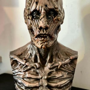 May include: A detailed bust of a character from the movie "Hellraiser." The sculpture is primarily brown with a textured, decaying appearance. The word "HELLRAISER" is displayed in white block letters at the base of the bust.