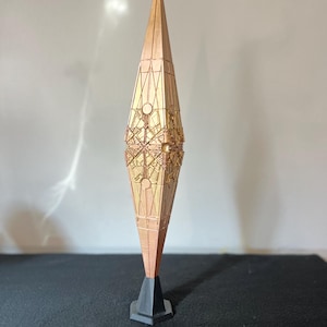 May include: A tall, copper-colored, spindle-shaped sculpture featuring intricate geometric patterns and circular motifs across its surface. The object tapers to a point at both the top and bottom and is mounted on a small, black, hexagonal base.