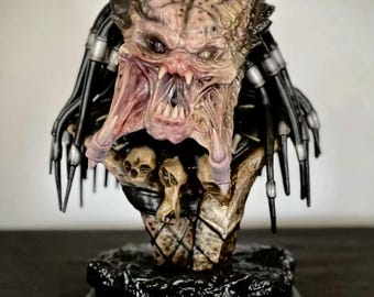 May include: A detailed bust of a Predator alien, with textured skin and a menacing expression. The sculpture features dreadlocks, mechanical elements, and a base with skulls. The alien's mouth is open, revealing sharp teeth.