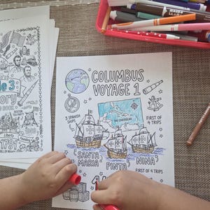 May include: A coloring book page with the text "Columbus Voyage 1" and illustrations of ships and a map of the Caribbean. A child is coloring the page with a red marker. A tray of markers is in the background.