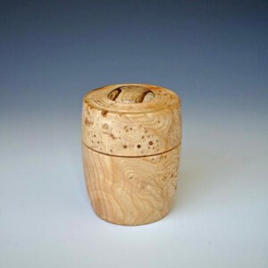 Könnte beinhalten: Eine kleine Holzkiste mit einem Deckel aus Maserholz. Die Kiste ist zylindrisch geformt und hat eine natürliche Holzoptik.