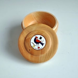 Puede incluir: Una caja de madera con una tapa redonda. La tapa tiene un diseño pintado de un pájaro con acentos rojos, azules y amarillos.