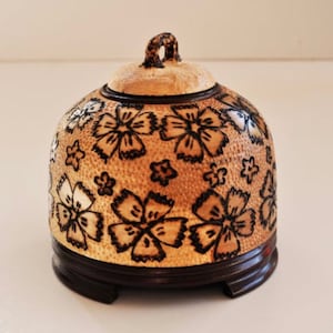Könnte beinhalten: Eine kleine, kuppelförmige Holzkiste mit einem Blumendesign. Die Kiste ist mit einem hellbraunen Hintergrund und schwarzen Blumenmustern bemalt. Die Kiste hat einen Deckel mit einem kleinen Griff.