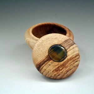 Könnte beinhalten: Eine Holzkiste mit Deckel. Der Deckel hat ein rundes, eingelegtes Stück Achat in der Mitte. Die Kiste ist aus hellbraunem Holz mit dunkelbraunen Streifen.