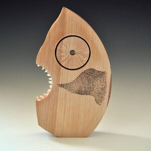 Puede incluir: Una escultura de madera de un pez con una gran boca abierta y dientes afilados. El pez tiene un ojo circular y un patr&oacute;n de puntos en su cuerpo.