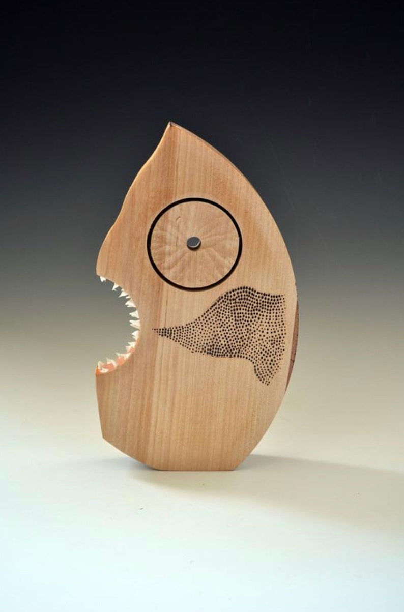 Puede incluir: Una escultura de madera de un pez con una gran boca abierta y dientes afilados. El pez tiene un ojo circular y un patr&oacute;n de puntos en su cuerpo.