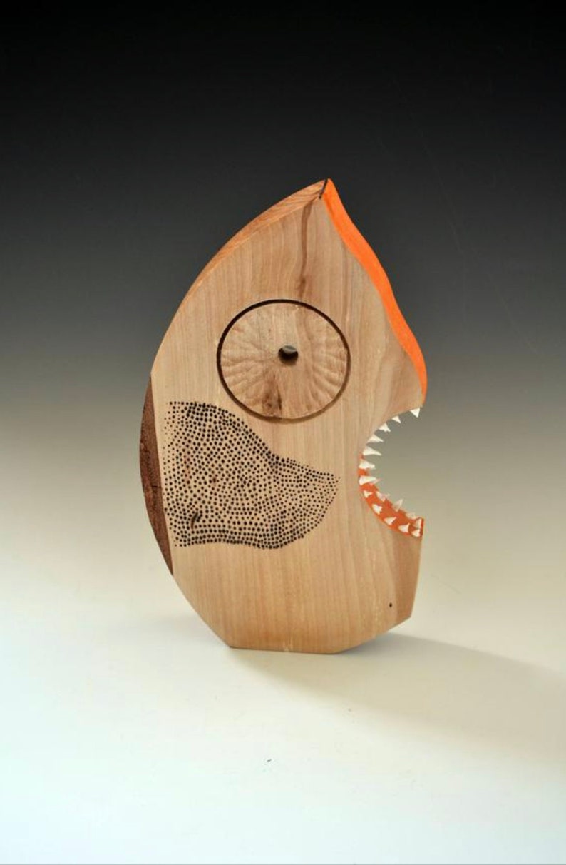 Puede incluir: Una escultura de madera de un pez con la boca abierta, pintada de naranja y blanco. El pez tiene un ojo circular y un patr&oacute;n de puntos negros y blancos en su cuerpo.