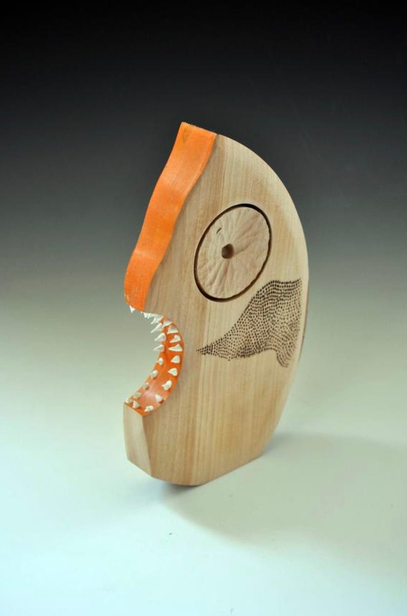 Puede incluir: Una escultura de madera de un pez con la boca pintada de naranja y un cuerpo con un patr&oacute;n en blanco y negro. El pez tiene un ojo grande y redondo y un conjunto de dientes afilados.