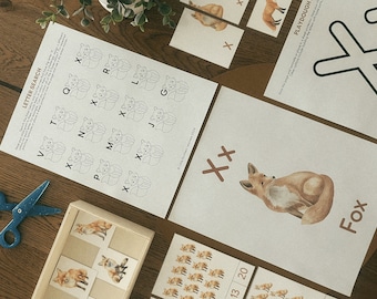 Op de afbeelding: Educatieve werkbladen met een letterzoektocht met vossenillustraties, een kleimat en flashcards. De bladen bevatten de letter X, het woord Fox en diverse vossenafbeeldingen. Een blauwe schaar ligt bij een houten dienblad met kaarten.