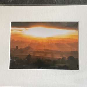 Puede incluir: Una fotografía de una salida del sol sobre un paisaje rural. El sol brilla a través de las nubes, creando un cálido resplandor dorado. El paisaje está cubierto por una ligera niebla, y se pueden ver árboles y campos en la distancia.