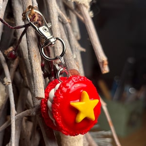 Peut inclure: Un porte-cl&eacute;s en forme de macaron rouge avec une &eacute;toile jaune, attach&eacute; &agrave; un fermoir argent&eacute;. Le charme est rond avec une garniture blanche et une &eacute;toile sur le devant. Le fermoir est attach&eacute; &agrave; un porte-cl&eacute;s argent&eacute;.
