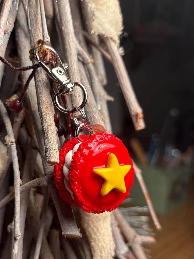 Peut inclure: Un porte-cl&eacute;s en forme de macaron rouge avec une &eacute;toile jaune, attach&eacute; &agrave; un fermoir argent&eacute;. Le charme est rond avec une garniture blanche et une &eacute;toile sur le devant. Le fermoir est attach&eacute; &agrave; un porte-cl&eacute;s argent&eacute;.