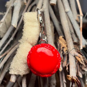 Peut inclure: Un charm rond rouge vif avec un bord d&eacute;coratif, attach&eacute; &agrave; un fermoir argent&eacute;. Le charm est suspendu devant un fond de brindilles claires et d'un petit objet duveteux cr&egrave;me.