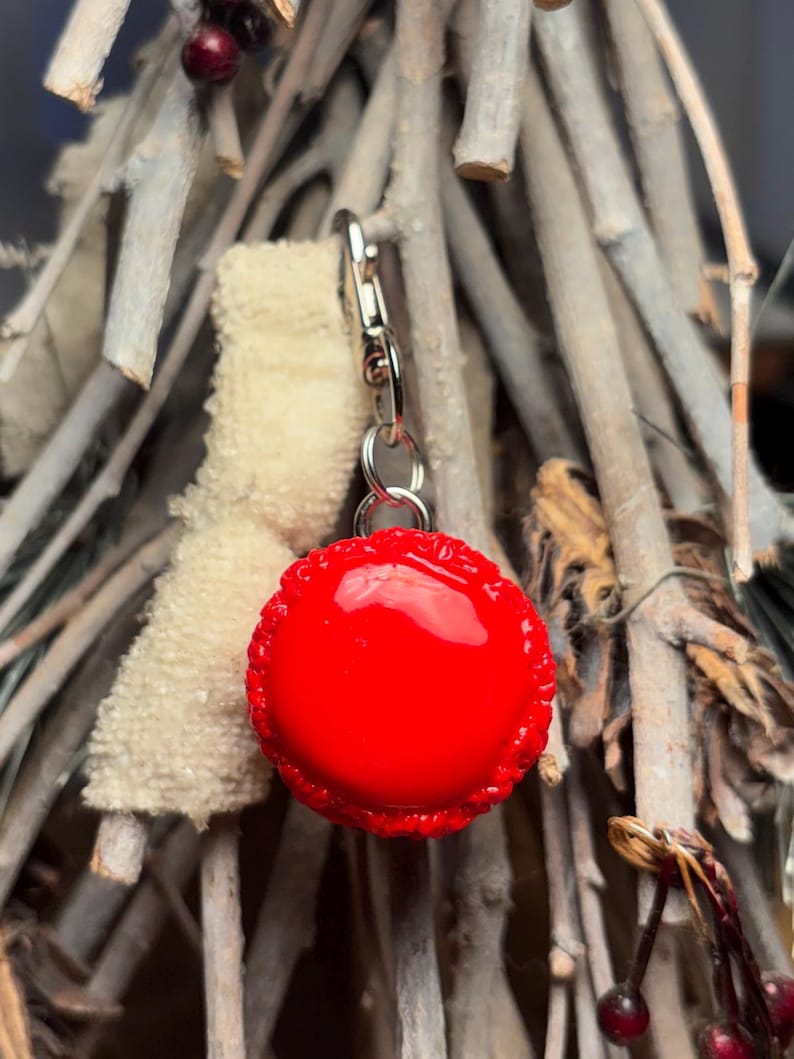 Peut inclure: Un charm rond rouge vif avec un bord d&eacute;coratif, attach&eacute; &agrave; un fermoir argent&eacute;. Le charm est suspendu devant un fond de brindilles claires et d'un petit objet duveteux cr&egrave;me.