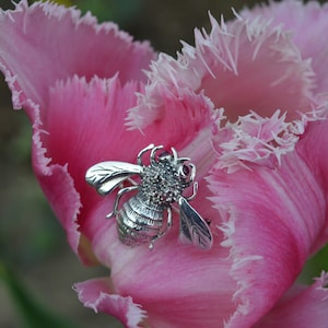 Puede incluir: Un broche de abeja de plata con una piedra preciosa de rubí sobre un tulipán rosa con flecos.