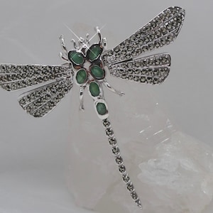 Puede incluir: Un broche de libélula de plata con detalles de piedras preciosas verdes. Las alas están cubiertas de pequeñas piedras brillantes.