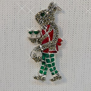 Op de afbeelding: Een zilveren broche met een grillige figuur die een groen-rood gepatroneerd pak draagt en een dienblad met twee groene glazen vasthoudt. De figuur is versierd met glinsterende marcasietstenen.