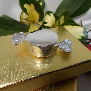 May include: A silver candy-shaped trinket box with a shiny, reflective surface and silver accents. The box sits atop a gold textured gift box, with green and yellow leaves and red berries in the background. A decorative item.