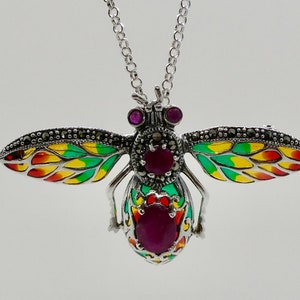 May include: A silver bee pendant necklace with colourful enamel wings in red, yellow, and green. The bee's body features ruby-coloured stones and marcasite accents. The necklace has a delicate chain.