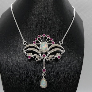 Peut inclure: Un collier en argent avec un pendentif en opale blanche entouré de pierres précieuses roses. Le pendentif est en forme d'éventail avec des détails complexes.