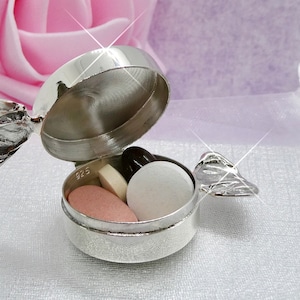 May include: A small, open silver pill box filled with various pills. The box is round with a hinged lid and decorative handles. The pills are different colors and shapes. The background is a soft pink and white.