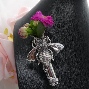 Puede incluir: Un broche de abeja de plata con un racimo de flores rosas en su cuerpo. La abeja está detallada con alas texturizadas y un cuerpo segmentado.