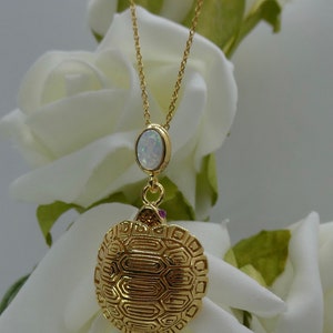 Puede incluir: Un collar de cadena de oro con un colgante de ópalo blanco y un dije de tortuga de oro. El dije de tortuga tiene un diseño de caparazón detallado.