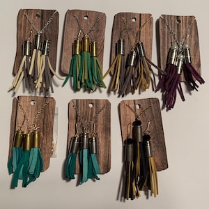 Op de afbeelding: Set van 8 paar oorbellen van patroonhulzen met leren kwastjes in verschillende kleuren, waaronder turquoise, groen, paars, zwart en beige. Elk paar wordt gepresenteerd op een houten kaart met een ketting.