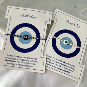 Könnte beinhalten: Zwei weiße Karten mit blauen und goldenen Glücksbringer-Armbändern. Die Karten tragen den Text "Evil Eye" und eine Beschreibung der Bedeutung des Glücksbringers.