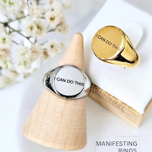May include: Two silver and gold signet rings with the words "I can do this" engraved on the top. The rings are displayed on wooden ring holders. The text "MANIFESTING RINGS" is displayed below the rings.