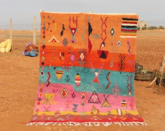 Könnte beinhalten: Ein farbenfroher, handgewebter Teppich mit geometrischen Mustern und einem Fransenrand. Der Teppich zeigt Bänder in Orange, Rot, Türkis und Rosa mit verschiedenen Formen und Symbolen in kontrastierenden Farben. Der Teppich wird im Freien präsentiert.