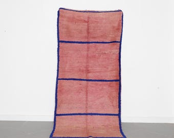 May include: A rectangular rug with a pink and red color scheme and blue horizontal stripes. The rug has a textured appearance and a fringed edge at the bottom. The rug is displayed against a white wall and a gray tiled floor.