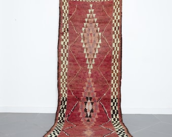 May include: A long, narrow red rug with a geometric pattern in shades of brown, white, and pink. The rug has a fringe on both ends.