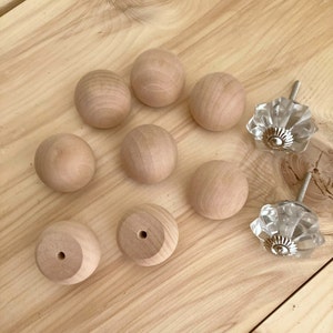 May include: A collection of wooden cabinet knobs and two clear glass crystal knobs with silver hardware. The wooden knobs are round and unfinished, showing the natural wood grain. The knobs are arranged on a light-colored wooden surface.