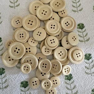 May include: A collection of various sized, light beige wooden buttons with four holes each. The buttons are scattered on a white fabric with a green floral pattern. The buttons are round and have a natural wood grain.
