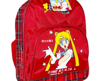 May include: Red backpack featuring a Sailor Moon graphic on the front. The backpack has a plaid pattern on the bottom and sides, with a matching tag. The graphic includes text in Japanese and English.