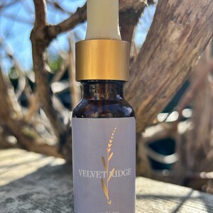 Puede incluir: Una pequeña botella de vidrio con una tapa dorada y un gotero. La botella tiene una etiqueta de color púrpura claro con el texto "VELVET RIDGE" y "15ml / .5oz" impreso en dorado.