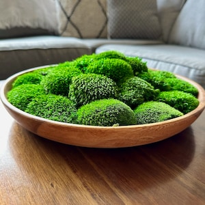 Op de afbeelding: Een houten kom gevuld met levendig groen mos, wat een natuurlijke, getextureerde presentatie creëert. De warme houttinten van de kom contrasteren met het heldere groen, waardoor het een decoratief accent voor een huis is. De kom staat op een houten tafel.