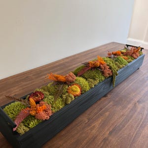 May include: A rectangular, black wooden planter box filled with vibrant green moss and dried flowers in shades of orange, red, and yellow. The arrangement is set on a wooden surface, creating a natural, decorative display.