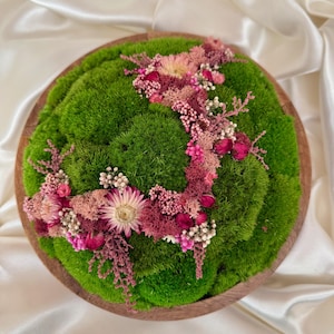 Könnte beinhalten: Ein runder Holztablett mit leuchtend grünem Moos, verziert mit einem zarten Arrangement aus getrockneten Blumen in Rosa-, Magenta- und Weißtönen. Das Blumendesign verläuft über das Moos und schafft ein natürliches Dekorationselement.
