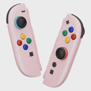 May include: Two pink game controllers with black accents. Each controller has a black joystick and colored buttons: red, yellow, green, and blue. The controllers are designed for gaming.