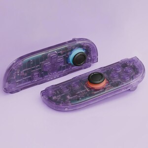 Puede incluir: Dos mandos de juego morados transparentes con componentes internos visibles. Uno tiene un anillo azul alrededor del joystick, el otro un anillo naranja. Ambos tienen joysticks y botones negros.