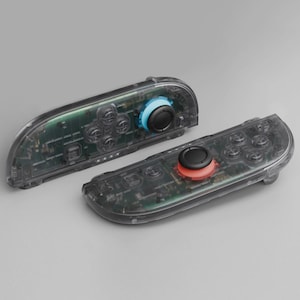 May include: Two transparent game controllers with visible internal components. One controller has a blue joystick and the other has a red joystick. The controllers have black buttons and a clear plastic shell.