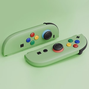 Op de afbeelding: Twee groene gamecontrollers met zwarte accenten en kleurrijke knoppen. De controllers hebben rode, gele, blauwe en groene knoppen. De joysticks zijn zwart met een rode ring.