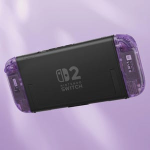 Puede incluir: Consola Nintendo Switch negra con mandos Joy-Con morados transparentes. La consola presenta el logotipo de Nintendo Switch y el n&uacute;mero "2". El fondo es de color morado suave.