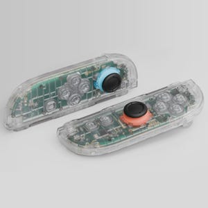 Könnte beinhalten: Zwei transparente Gamecontroller mit sichtbarer interner Schaltung. Ein Controller hat einen blauen Joystick und der andere einen orangefarbenen Joystick. Beide Controller haben schwarze Tasten und ein transparentes Kunststoffgehäuse.